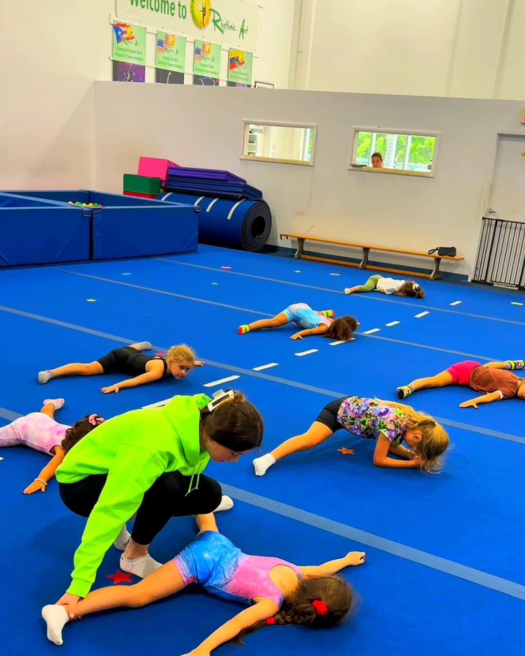 A coach kneels to guide a girl's stretch while the class stretches on the blue floor at Rhythmic Art